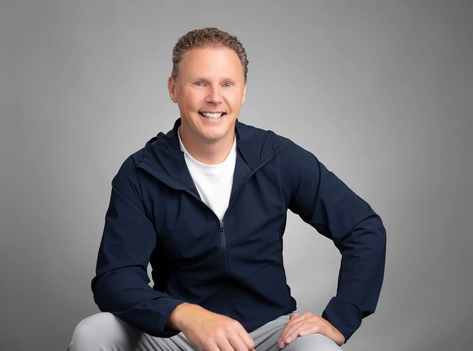 Chad Richardson seated, one hand resting on his knee, smiling at the camera in a navy jacket over a white tee.