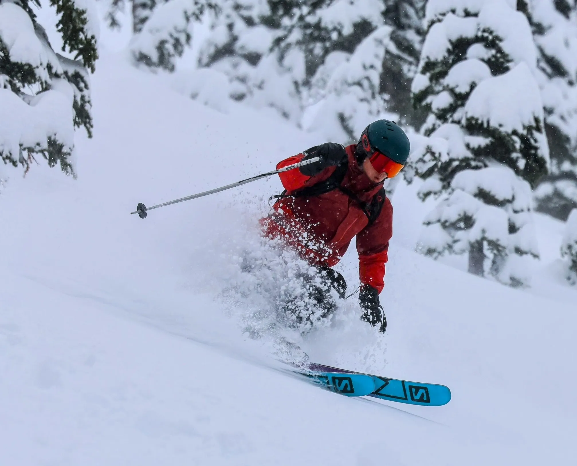 A skier in red carving through deep powder past snow-covered trees.