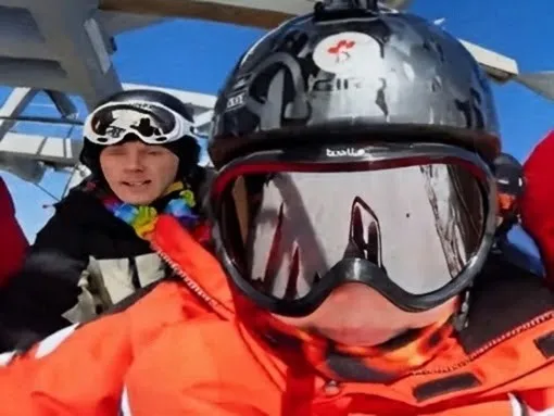 A skier in an orange bib riding a chairlift, Chad alongside in a helmet and goggles.