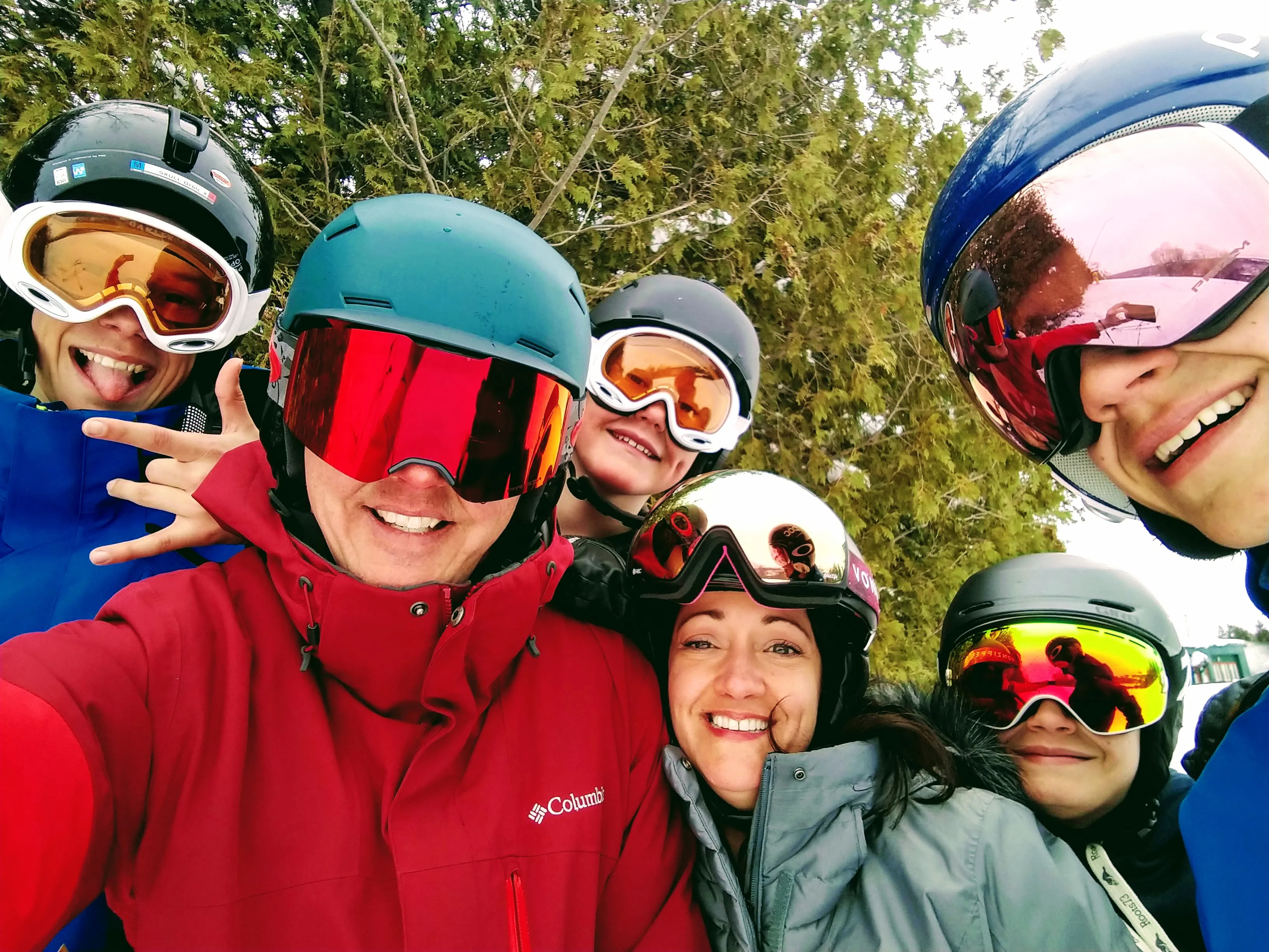 A close selfie of 6 of the family in ski helmets and goggles, all grinning.