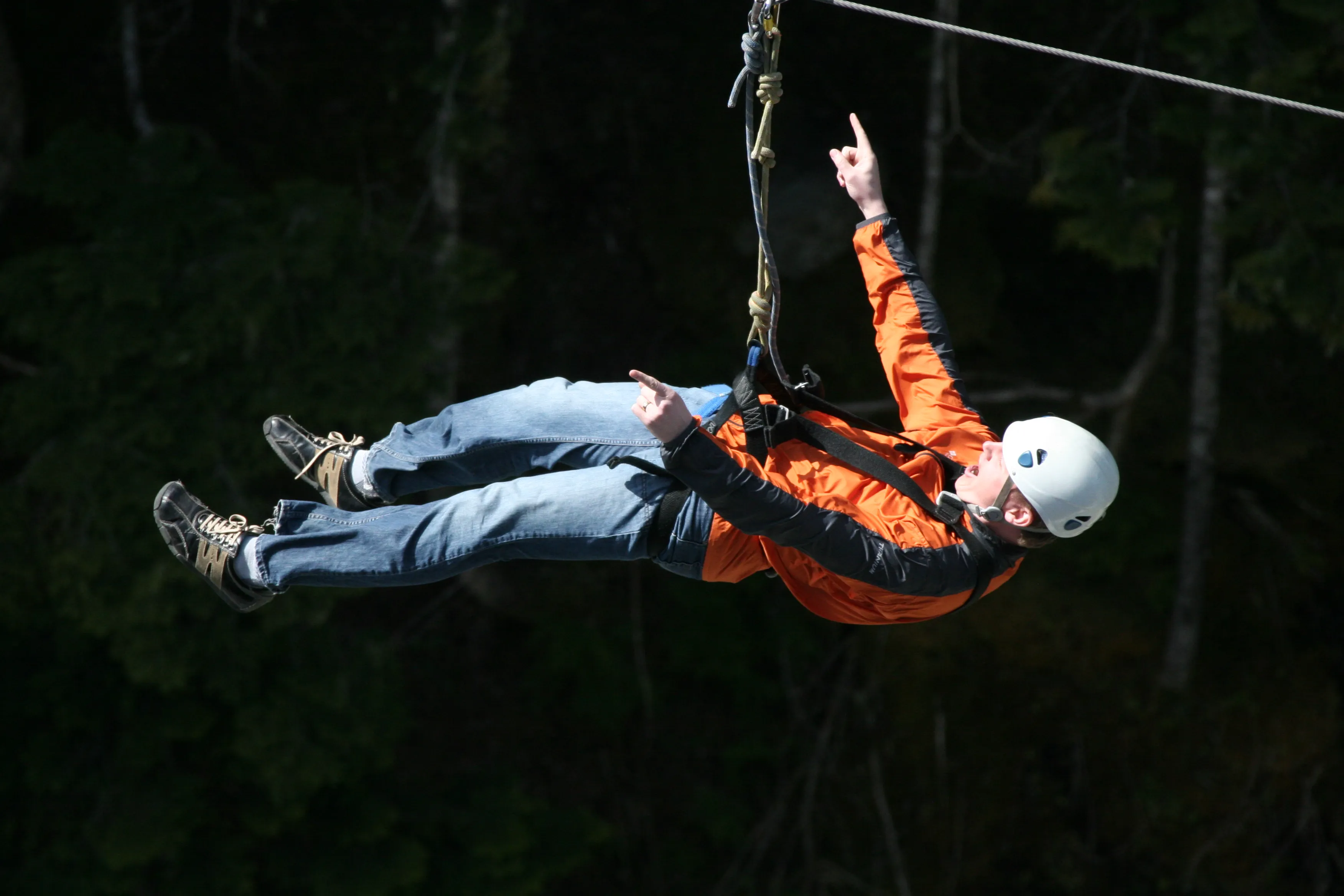 Chad mid-ride on a zip line, harnessed and pointing both hands skyward, forest behind him.