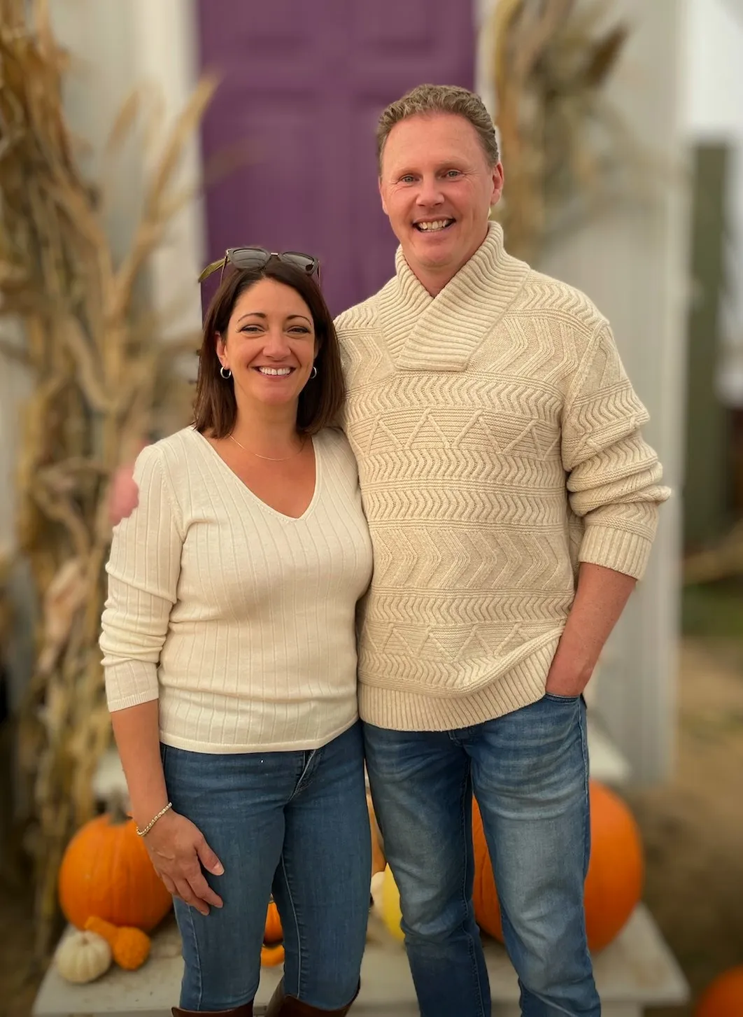 Chad and Liz in cream sweaters beside a purple door, pumpkins and cornstalks around them.