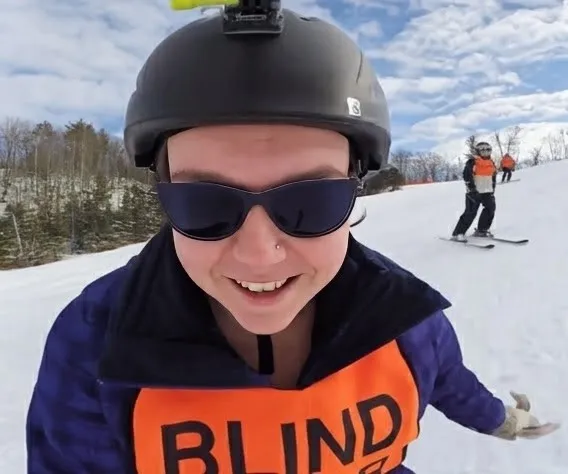 A skier in a BLIND SKIER bib smiling on the slope, another skier behind.